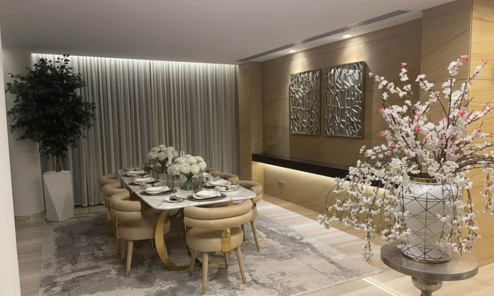 A sophisticated luxury interior design dining room. The room features a marble dining table with a sleek gold-toned sculptural base. The table is surrounded by eight plush, upholstered dining chairs in a soft beige color. On the table, there is a formal place setting with white dinnerware and a centerpiece of two large vases filled with white roses. The walls are a mix of light-colored wood paneling with subtle lighting, and a feature wall with two modern, three-dimensional metallic art pieces. To the left, a large potted plant stands next to sheer white curtains.