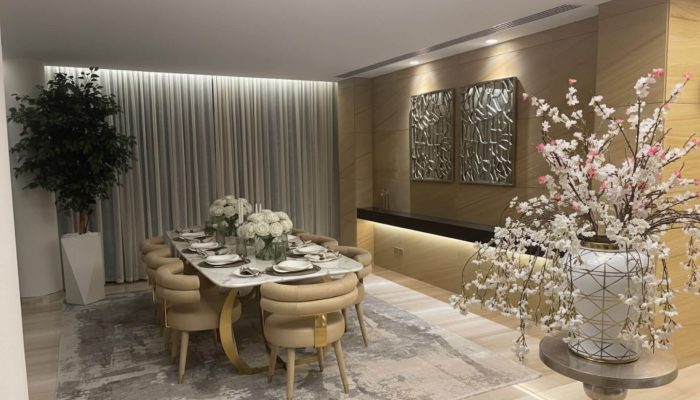 An elegant and modern dining room featuring a long table, upholstered chairs, a tiered chandelier, and a decorative mirror wall