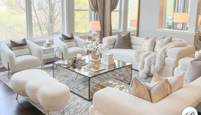 A luxurious and bright living room is decorated in a neutral, monochrome palette of white, cream, and beige. The space features two cream-colored bouclé slipper chairs and a matching textured ottoman. A large, plush sectional sofa is covered with throw pillows in varying textures and shades of beige and a soft, fluffy blanket. The centerpiece is a large, square mirrored coffee table with gold accents, topped with decorative items like vases of white flowers, candles, and books. The room is anchored by a patterned area rug and has large windows that let in ample natural light. A large, ornate gold-framed mirror leans against the far wall.