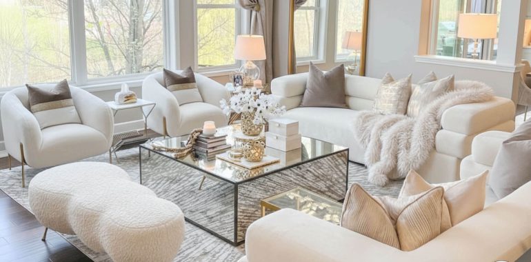 A luxurious and bright living room is decorated in a neutral, monochrome palette of white, cream, and beige. The space features two cream-colored bouclé slipper chairs and a matching textured ottoman. A large, plush sectional sofa is covered with throw pillows in varying textures and shades of beige and a soft, fluffy blanket. The centerpiece is a large, square mirrored coffee table with gold accents, topped with decorative items like vases of white flowers, candles, and books. The room is anchored by a patterned area rug and has large windows that let in ample natural light. A large, ornate gold-framed mirror leans against the far wall.