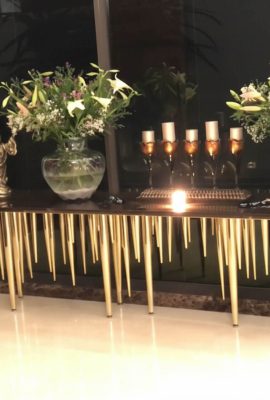 A stunning console table for a luxury interior design entryway or living space. The table features a long, dark top and is supported by a unique, striking base of numerous thin, tapered gold metal legs that resemble icicles or rain. The table is styled with two large glass vases filled with fresh flowers, a set of lit candles in elegant holders, and a small golden statue. The polished floor reflects the ambient lighting, and large windows are visible in the background.